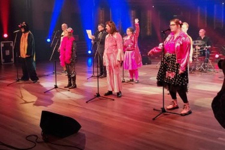 Op een podium staan mensen in mooie kostuums achter microfoons. Helemaal achterin zit een drummer.